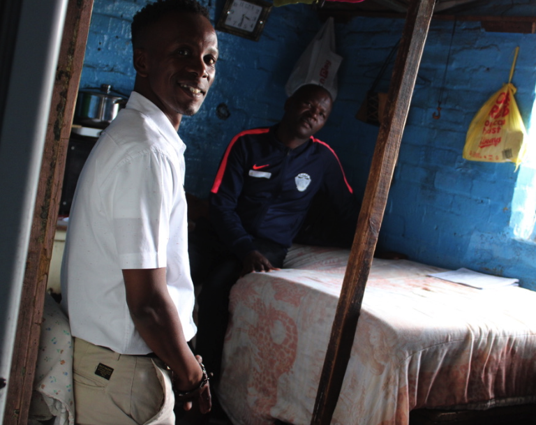 Ndoda Mbombo sits on the bed he shares with his wife and five year old. Our guide Bulelani Dondashe in foreground