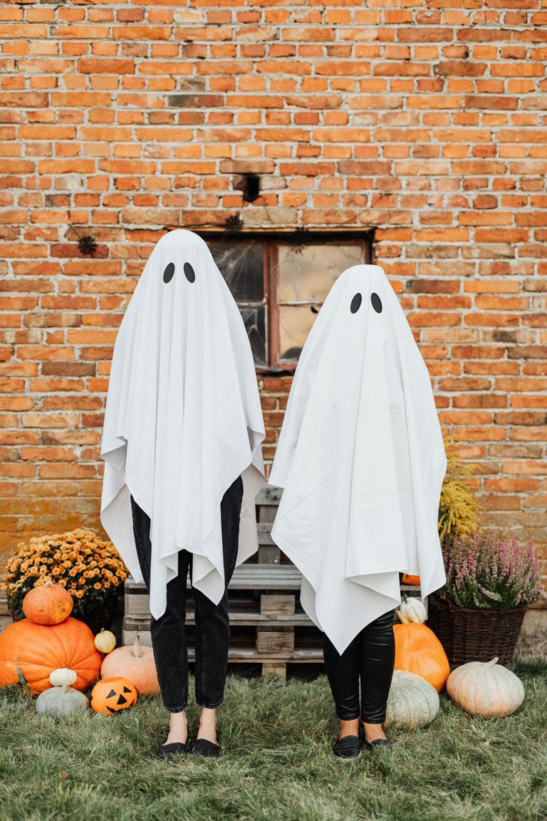 Two people in ghost costumes stand in front of a rustic brick wall surrounded by pumpkins, embodying Halloween spirit.