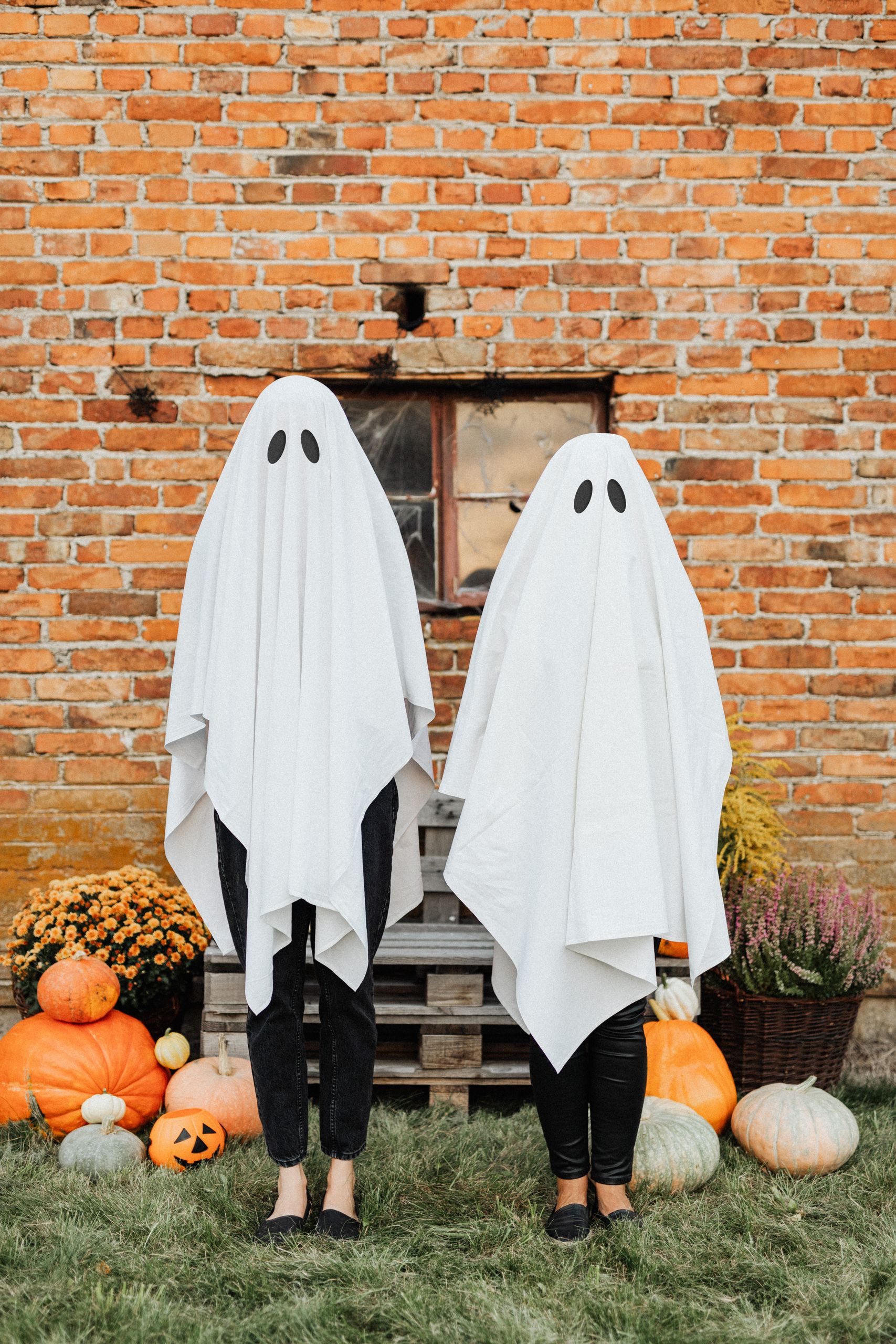 Two people in ghost costumes stand in front of a rustic brick wall surrounded by pumpkins, embodying Halloween spirit.