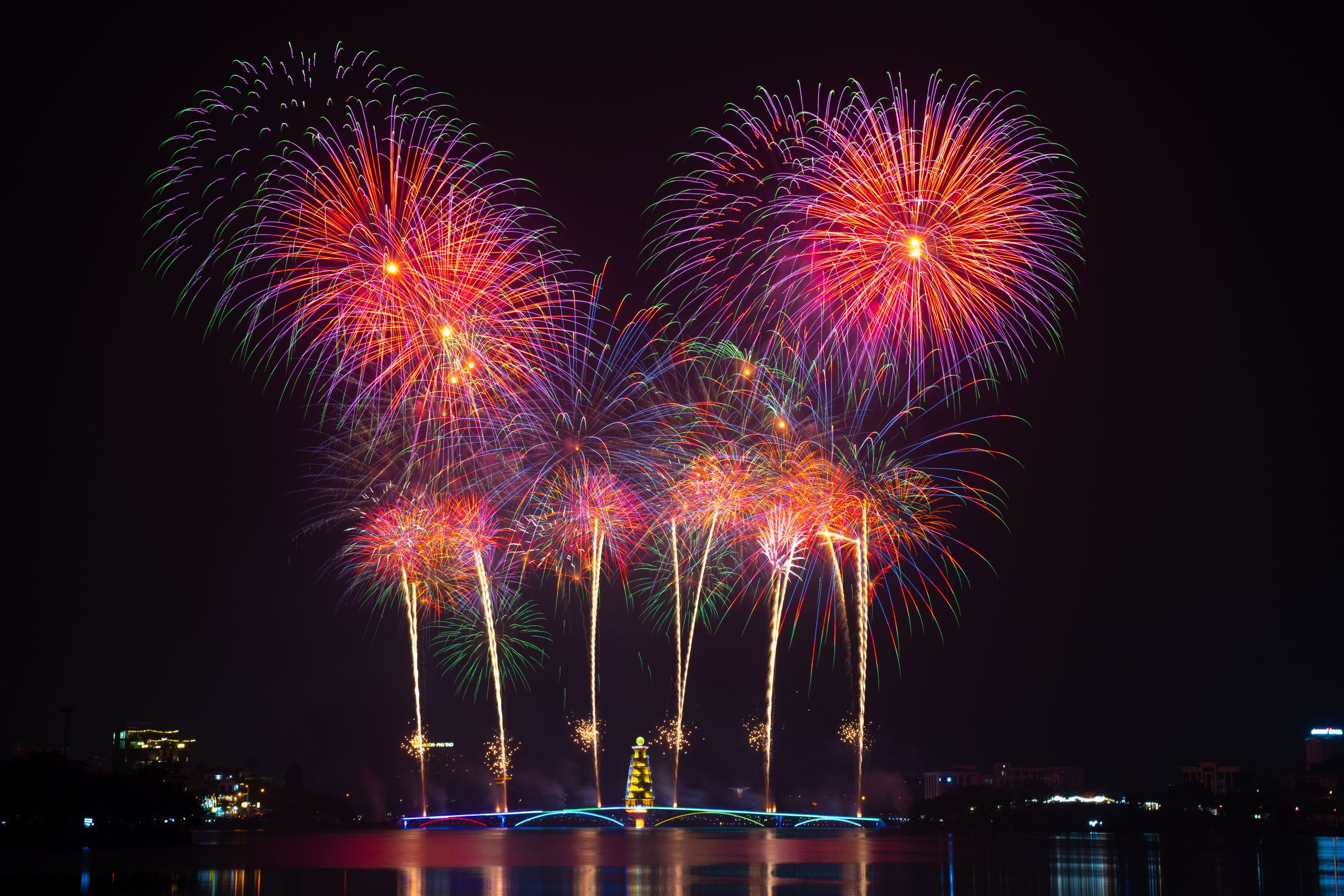 Vivid fireworks burst over a peaceful water scene, lighting up the night sky with colorful reflections.