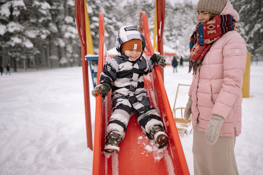 A child in winter attire slides down a playground slide with a mother watching in a snowy park.